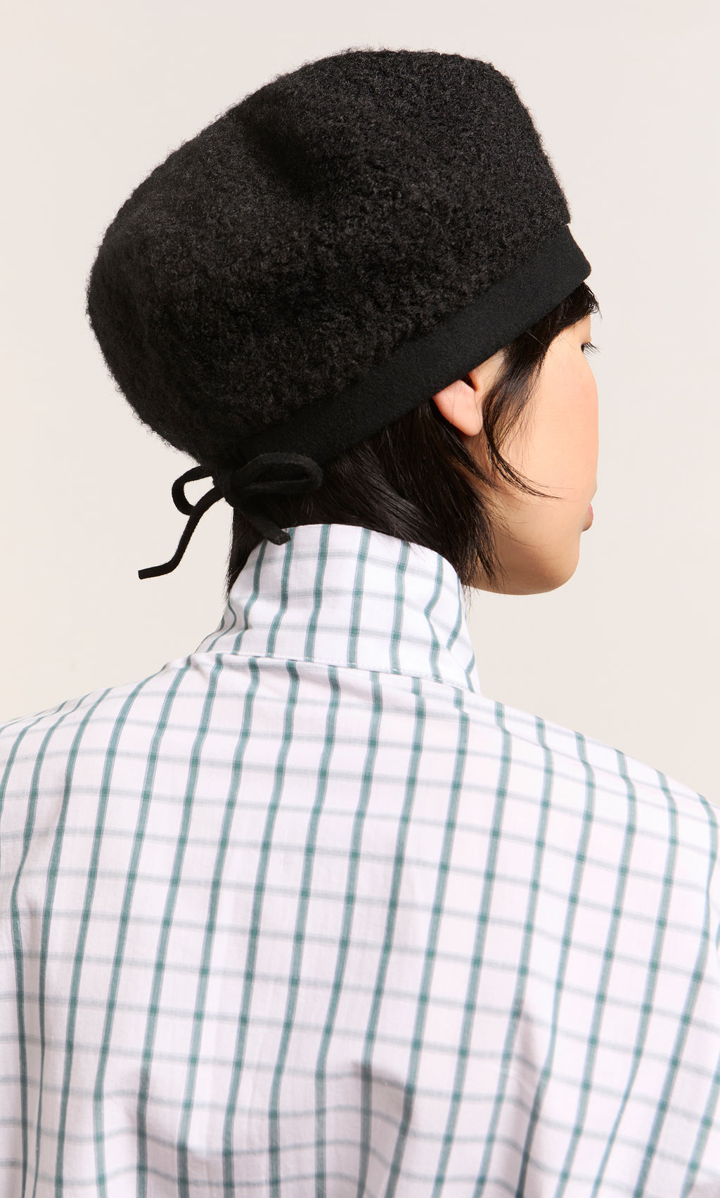 Plümo  Person wearing the Perinne Beret in black boucle, adjustable fit, with a plaid shirt, shown from behind on light background.