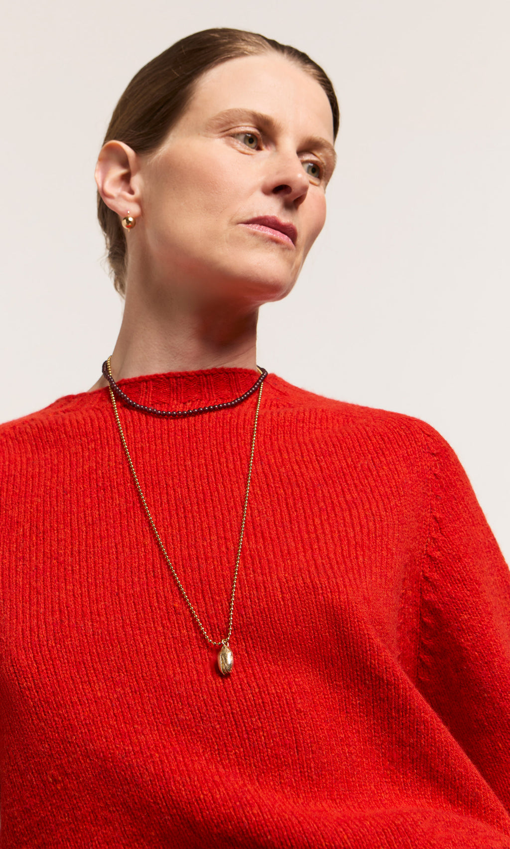 Plümo  Woman in a red sweater wearing the Pistachio Pendant and small gold hoops, gazing slightly to the side.