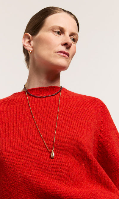Plümo  Woman in a red sweater wearing the Pistachio Pendant and small gold hoops, gazing slightly to the side.