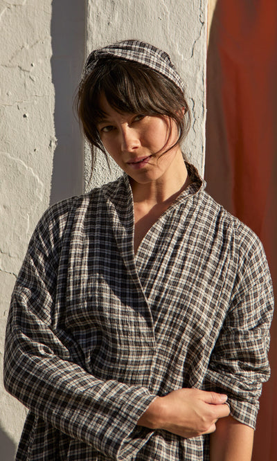 Plümo- Woman wearing a plaid headband stands by a white textured wall, looking at the camera in warm sunlight.