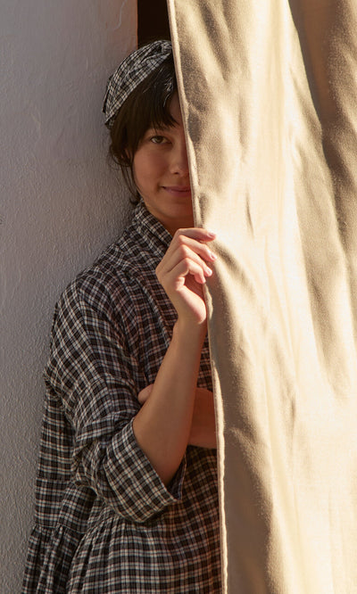 Plümo- Smiling in soft sunlight, a person peeks out with a Plaid Headband partly hidden by a curtain.
