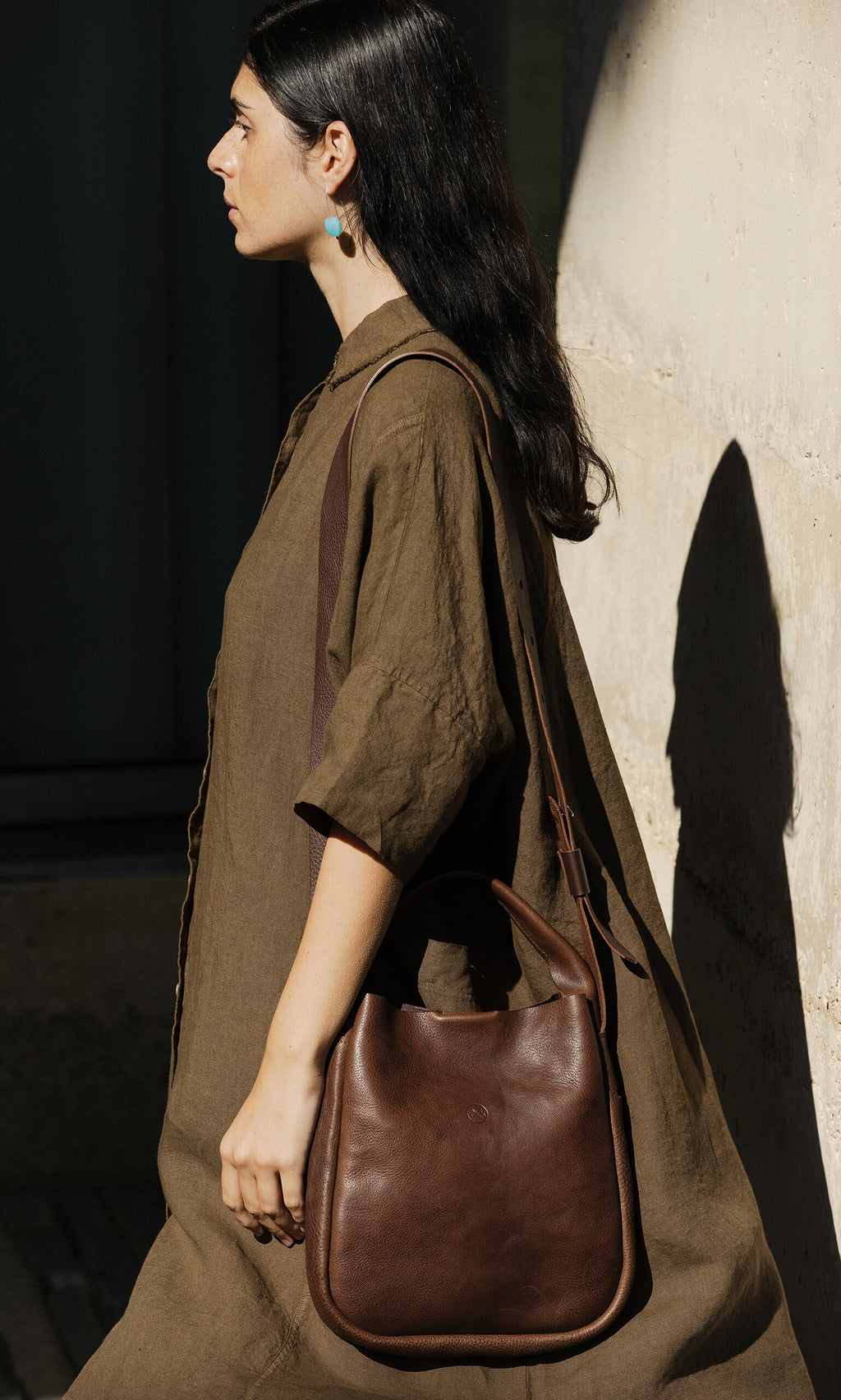 Plümo- Woman in a loose brown dress carries the Otto Leather Bag, walking beside a sunlit wall.