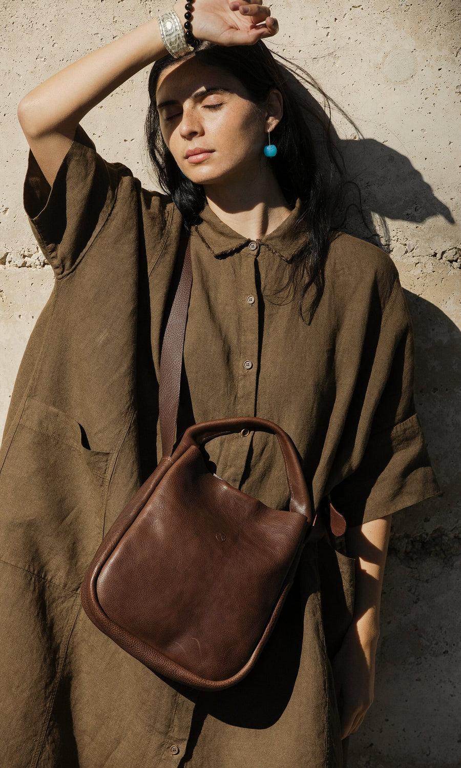 Plümo- Woman in a loose brown dress and blue earrings poses with the Otto Leather Bag by a sunlit concrete wall.