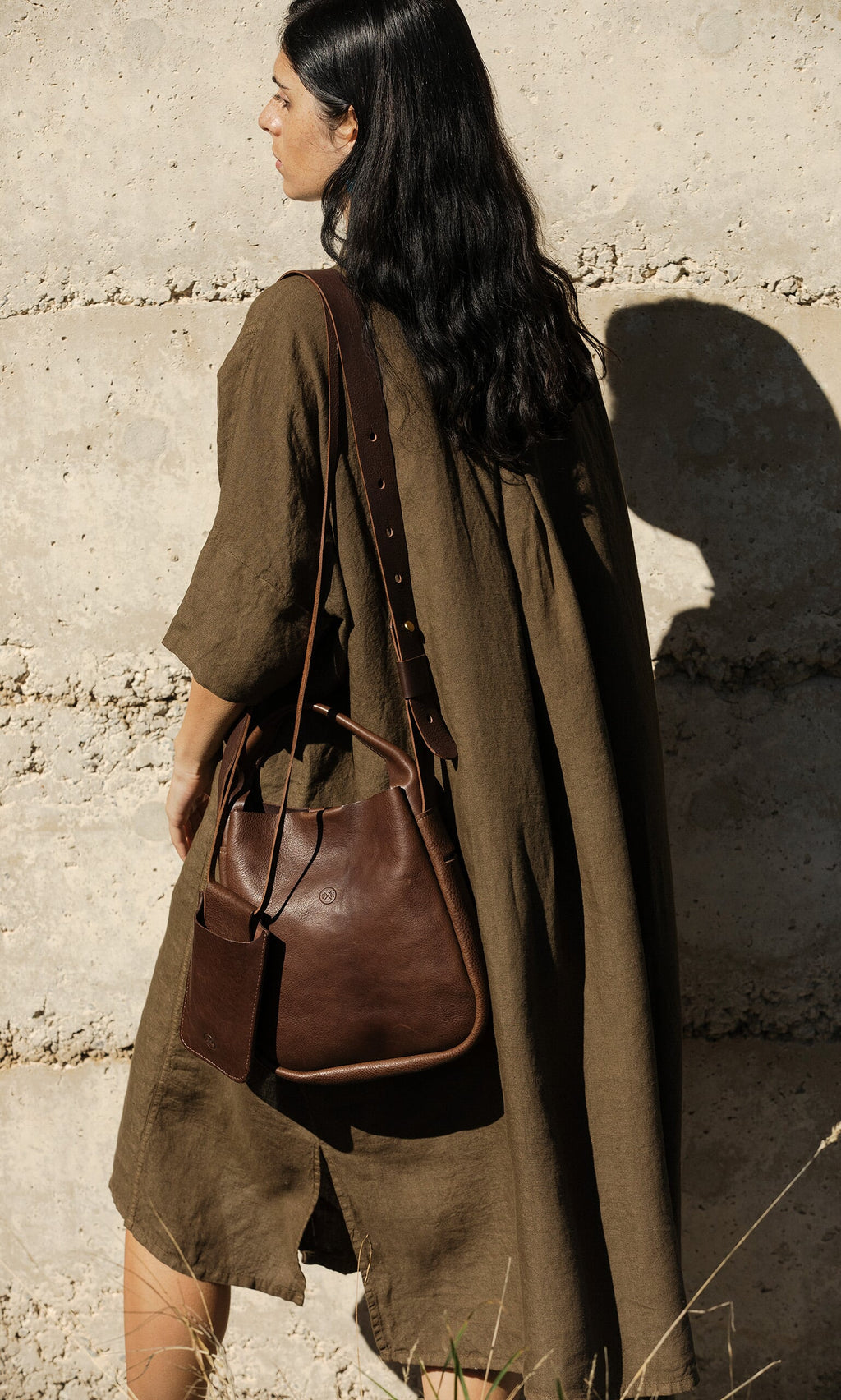Plümo- Woman with long dark hair in a brown dress carries the Otto Leather Bag with crossbody strap by a textured wall.