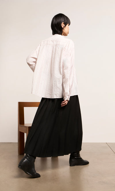Plümo  Person with short dark hair in an Ejaz Shirt stands by a wooden chair on a light floor.