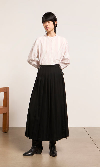 Plümo  Person wears the white Ejaz Shirt with stripes and a long black pleated skirt, standing indoors with hands behind back.