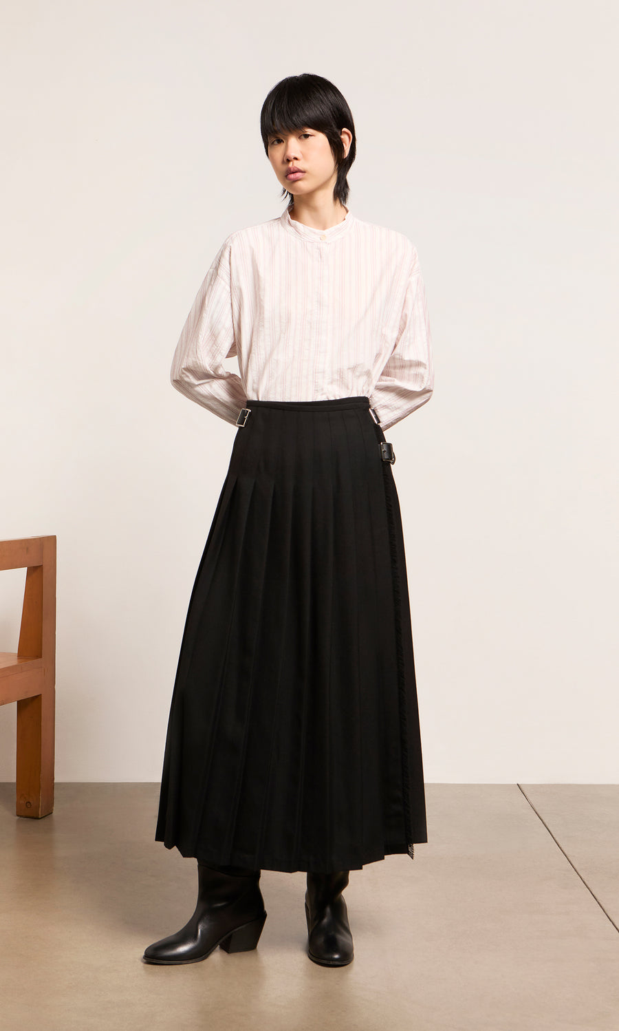 Plümo  Person in a white striped shirt and Storm Kilt stands indoors with hands behind their back.