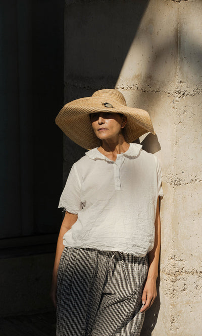 Plümo- Woman in a wide-brimmed hat wears the Renville Shirt, standing by a sunlit stone wall, partly in shadow.