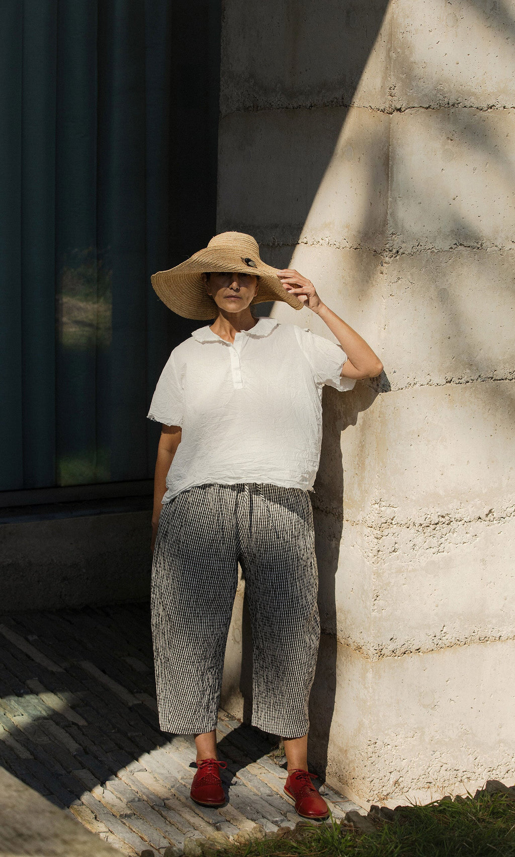Plümo- Woman in Serena Straw Hat, white shirt, striped pants, and red shoes stands by a sunlit concrete wall.