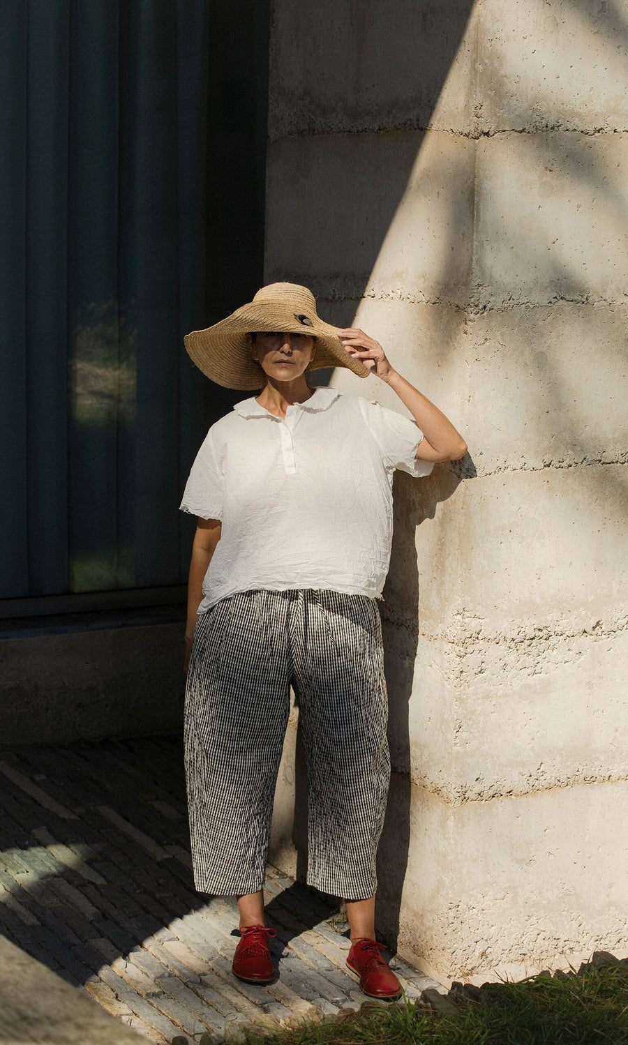 Plümo- Woman in Serena Straw Hat, white shirt, striped pants, and red shoes stands by a sunlit concrete wall.