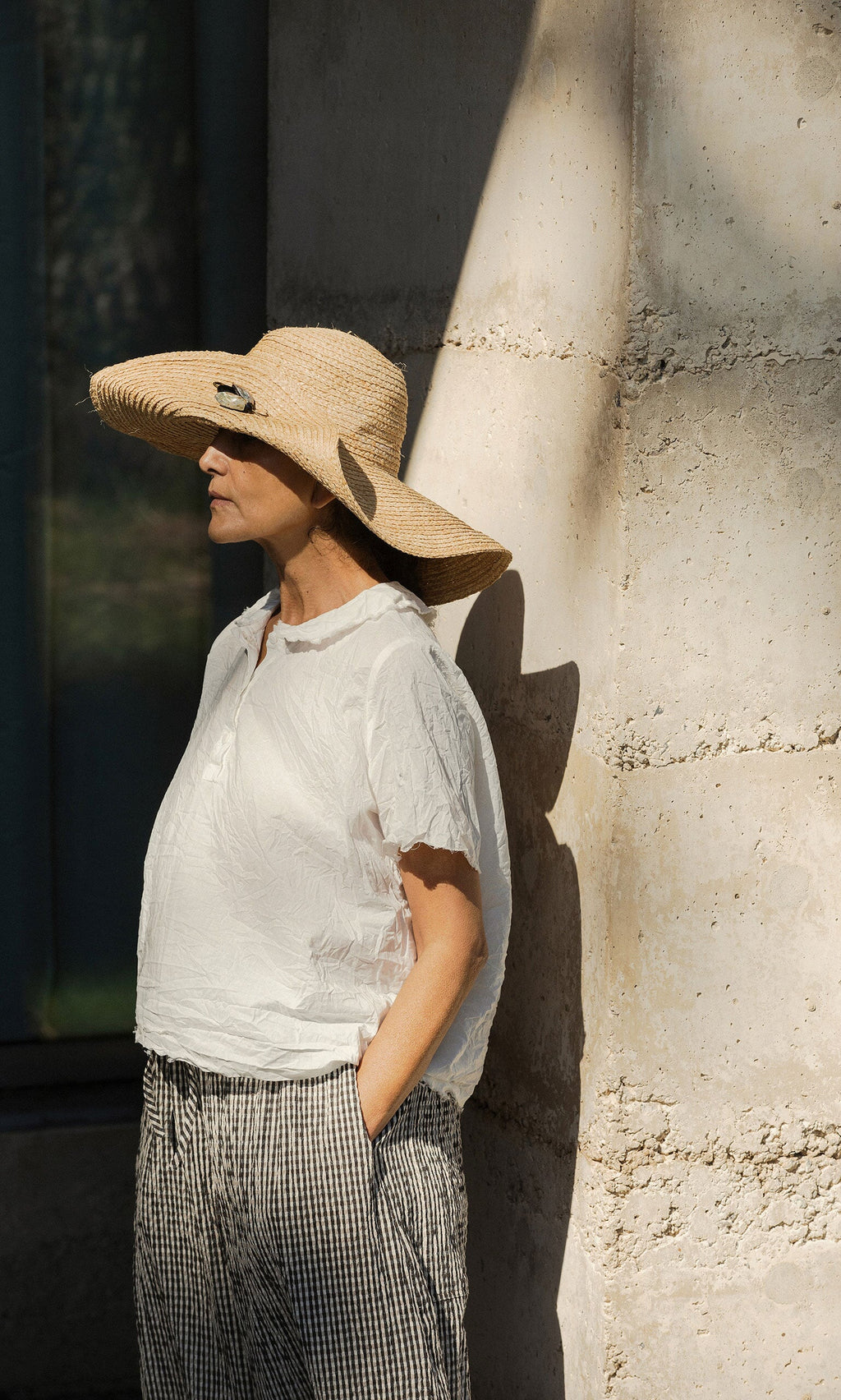 Plümo- Woman in a Serena Straw Hat and white blouse stands against a sunlit stone wall, hands in pockets.