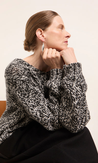 Plümo  Woman wearing the Rhian Wool Jumper sits sideways, chin on hands, appearing thoughtful in a sustainably-made sweater.