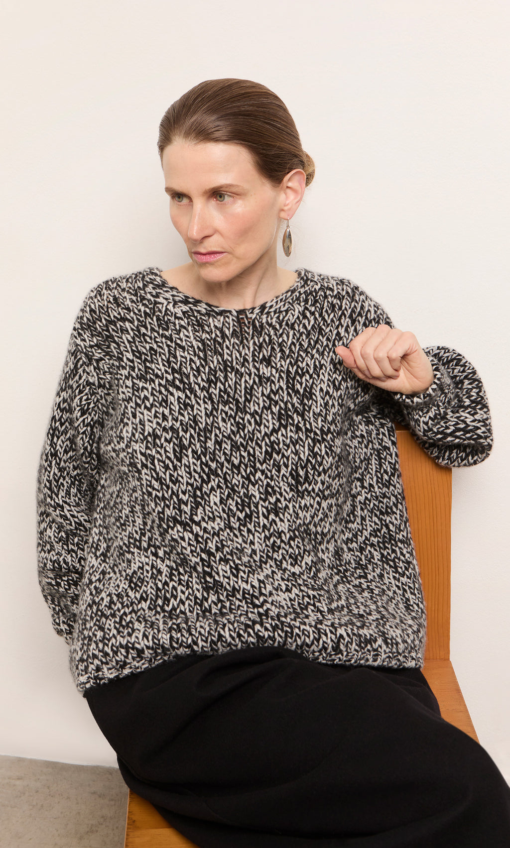 Plümo  Woman with brown hair in a bun wears the Rhian Wool Jumper, sitting on a wooden chair against a white wall.