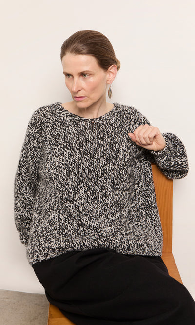 Plümo  Woman with brown hair in a bun wears the Rhian Wool Jumper, sitting on a wooden chair against a white wall.