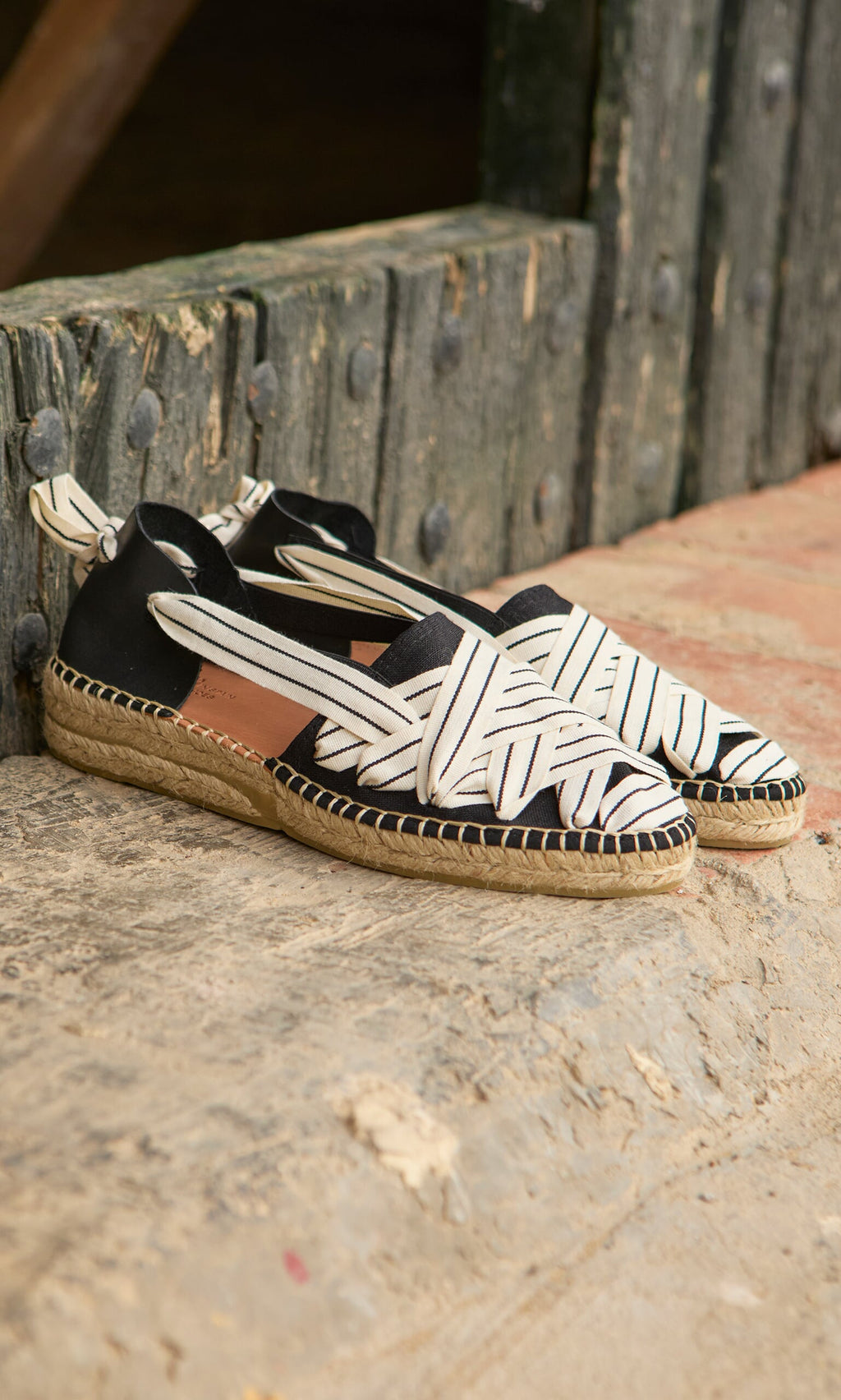 Plümo- Ribbon Espadrilles in black with white crisscross laces and leather heel, displayed on stone and brick.