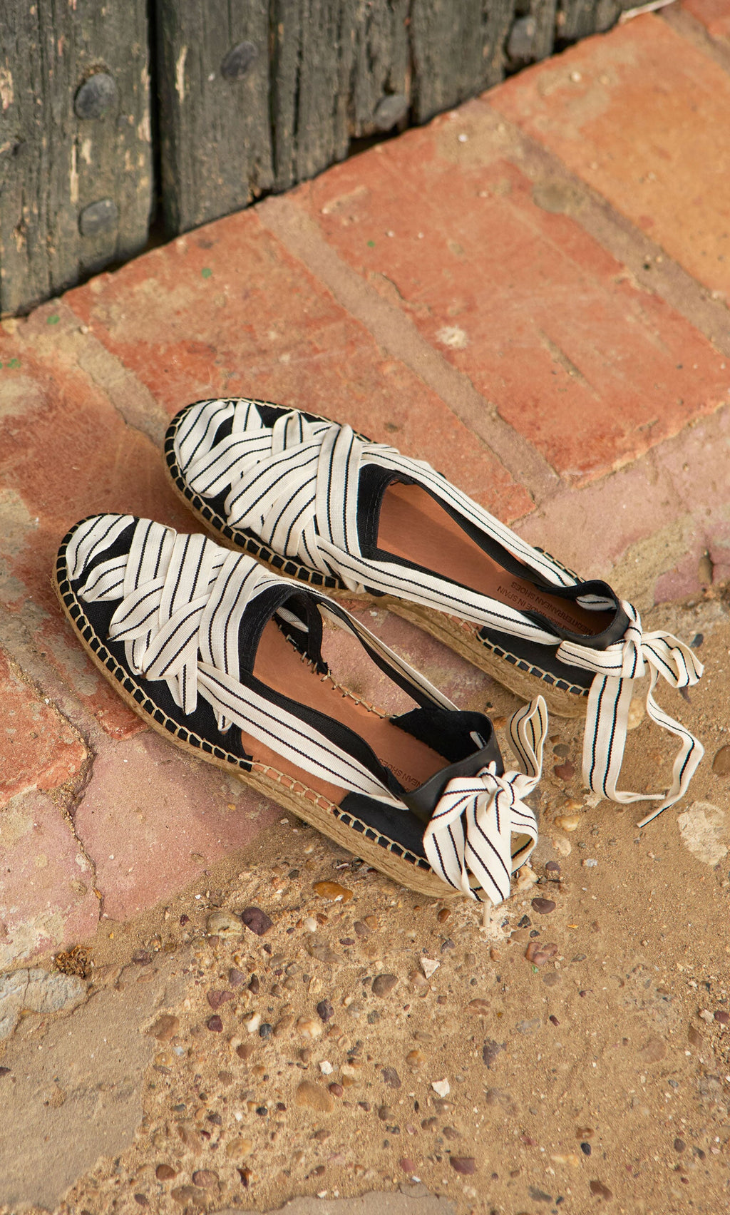 Plümo- Ribbon Espadrilles with bow ties and leather heels rest on brick and stone by a weathered wooden door.