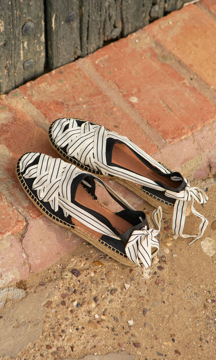 Plümo- Ribbon Espadrilles with bow ties and leather heels rest on brick and stone by a weathered wooden door.