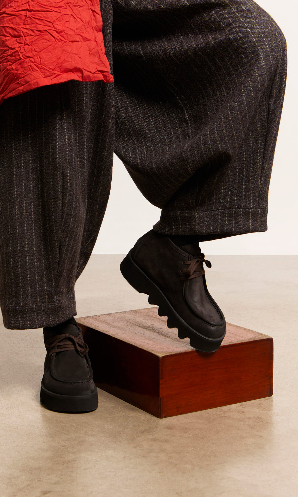 Plümo  Person in Rolla Boots with platform soles steps on a wooden block. Wide pinstripe pants and red garment partly visible.