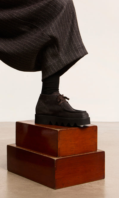 Plümo  Someone in dark pants steps onto stacked wooden boxes, wearing Rolla Boots—handcrafted Italian mocha suede moccasins.