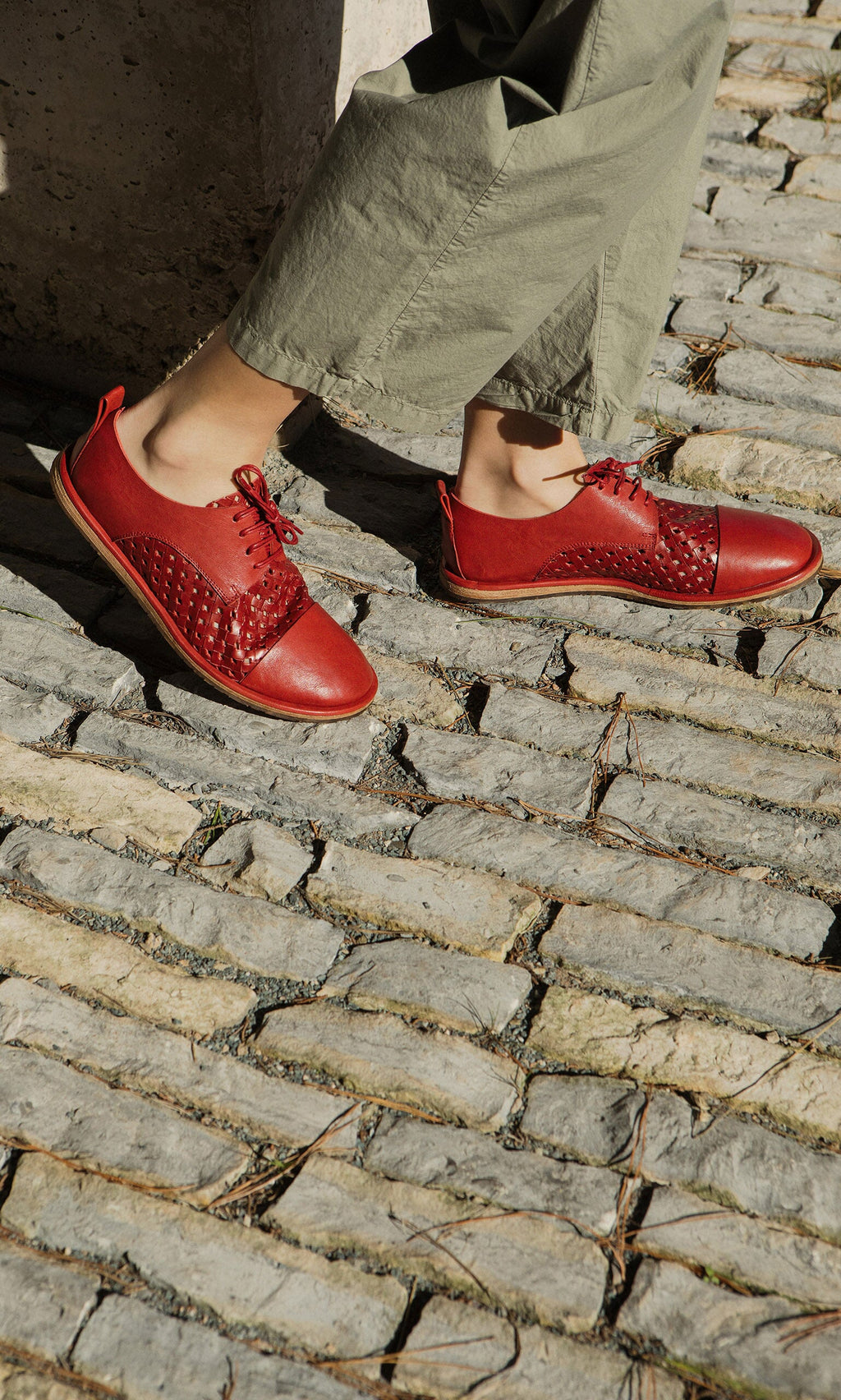 Plümo- Someone stands on a cobblestone street in sunlight, wearing olive pants and Magui Brogues in bright red Italian leather.