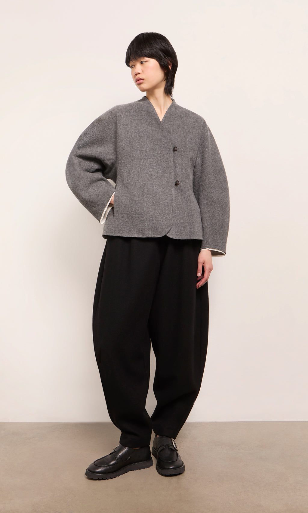 Plümo  Model wears Circle Pants - Boiled Black and a gray buttoned jacket, standing against a plain background.
