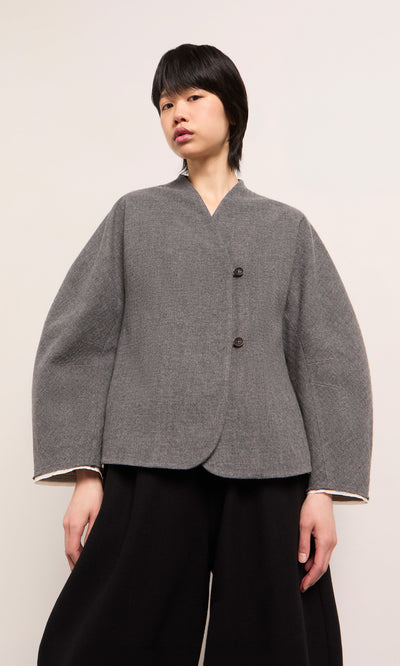 Plümo  Person wearing a grey Rostock Jacket with two buttons and wide black pants, standing against a plain background.