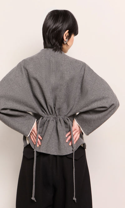 Plümo  Person in a grey Rostock Jacket by Plümo Studio, peplum hem, hands on hips, facing away from the camera.
