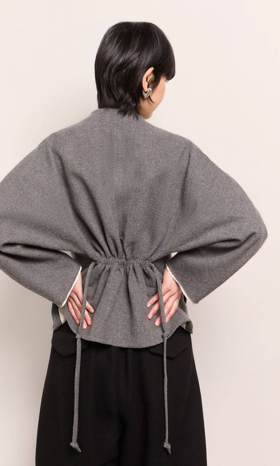Plümo  Person in a grey Rostock Jacket by Plümo Studio, peplum hem, hands on hips, facing away from the camera.