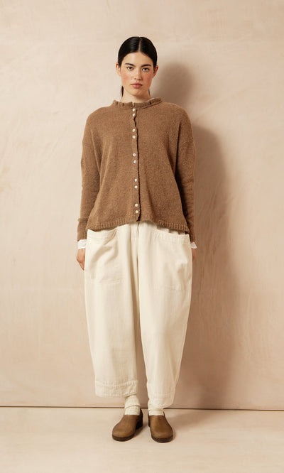 Plümo- Woman in Rutland Cardigan with rib-knit cuffs, cream wide-leg pants, and brown shoes against a beige wall.