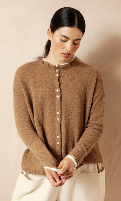 Plümo- Woman with dark hair wears the ultra-soft Rutland Cardigan with rib-knit cuffs and cream pants, looking down.