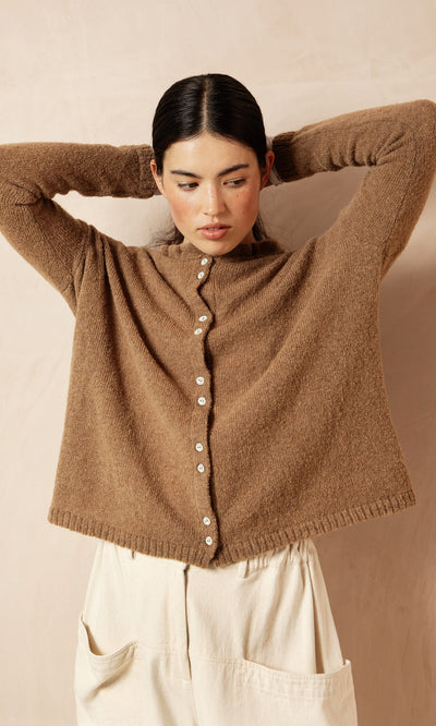 Plümo- Woman in the Rutland Cardigan with rib-knit cuffs and cream pants stands with arms raised against a beige background.