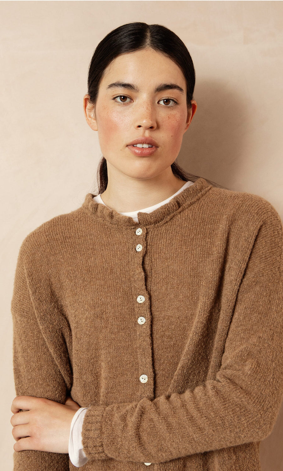 Plümo- Woman with dark hair in a low ponytail wears the Rutland Cardigan with rib-knit cuffs, against a beige background.