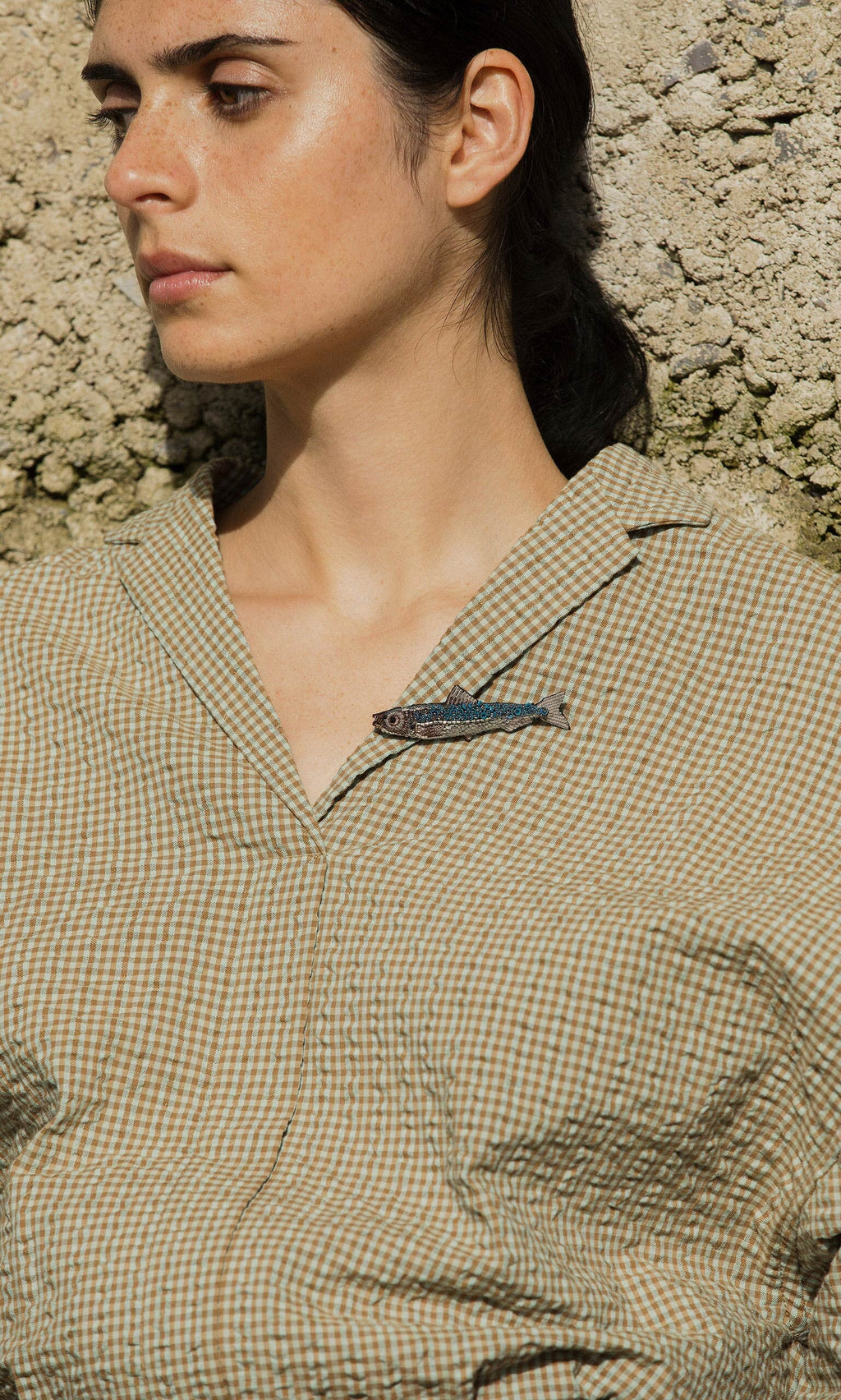 Plümo- A person in a checked shirt wears the Sardine Brooch, standing against a textured wall.