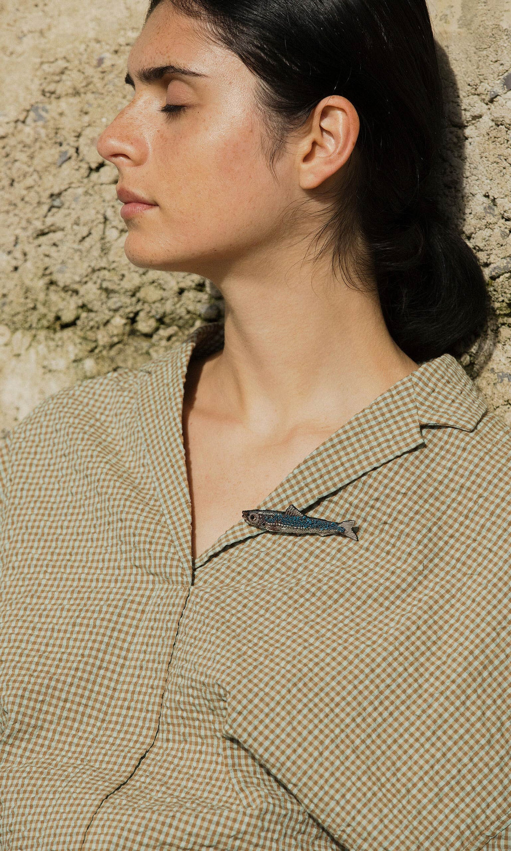 Plümo- A woman with closed eyes leans against a stone wall; a Sardine Brooch peeks from her shirt collar.