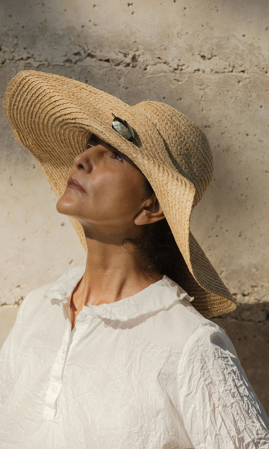 Plümo- Woman in the Serena Straw Hat and a white blouse stands against a textured stone wall in sunlight.