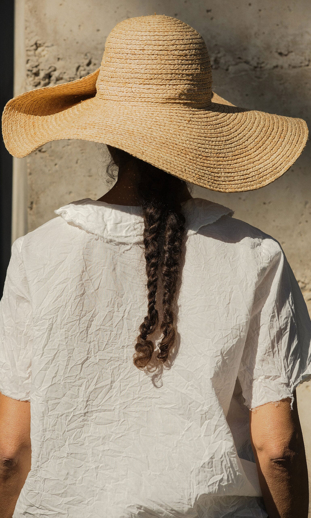 Plümo- A woman with braided hair wears the Serena Straw Hat and a crinkled white shirt, seen from behind.