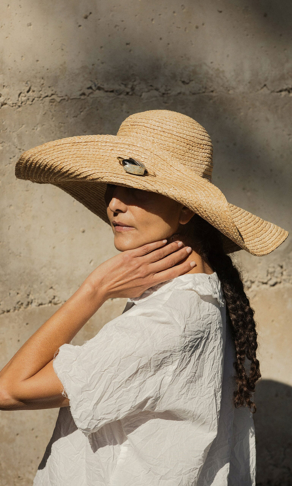 Plümo- Woman in a Serena Straw Hat and white blouse stands in sunlight, hand on neck, against a shadowed background.