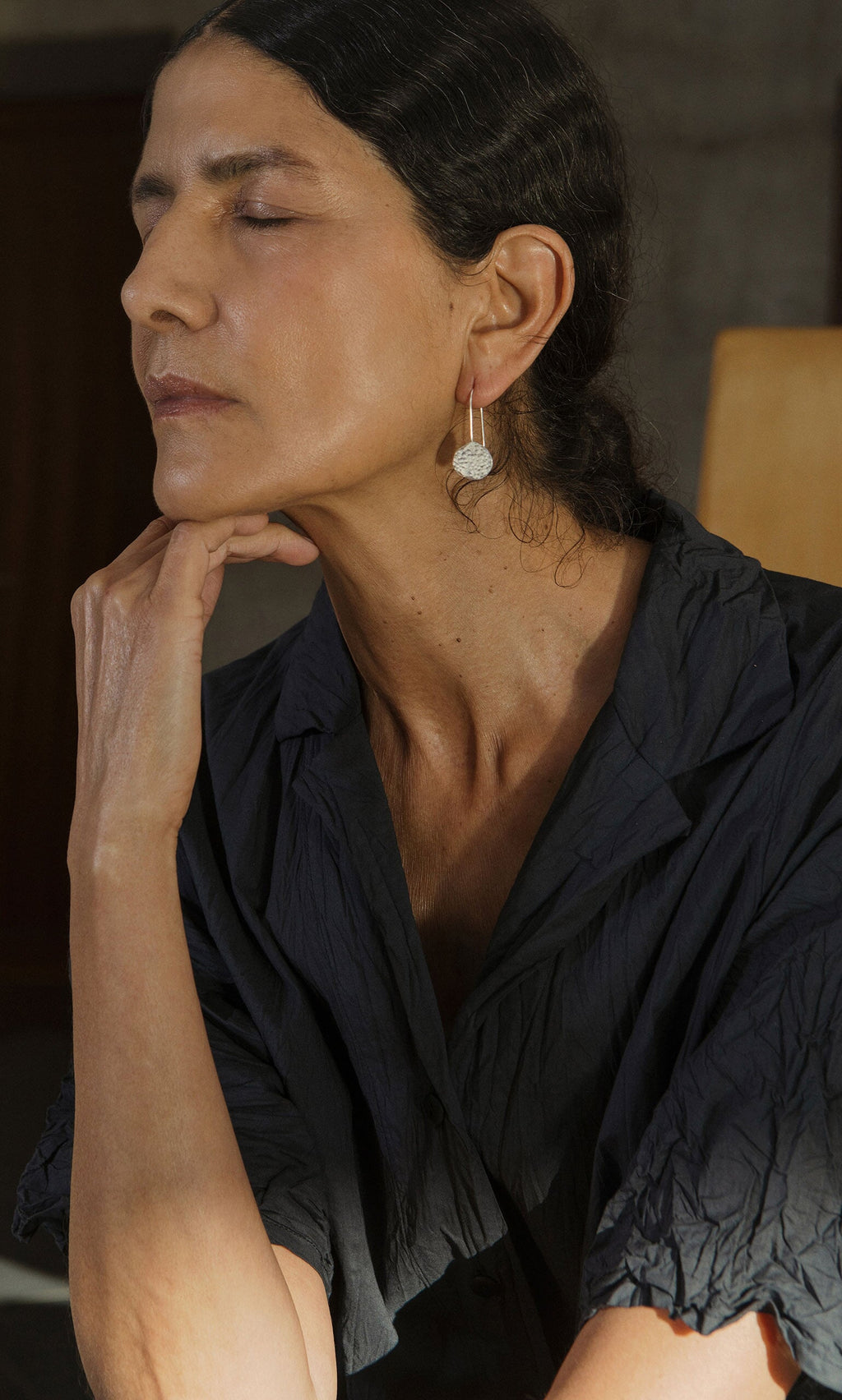 Plümo- A woman with closed eyes rests her chin on her hand, wearing Shirley Earrings and a dark crinkled shirt.