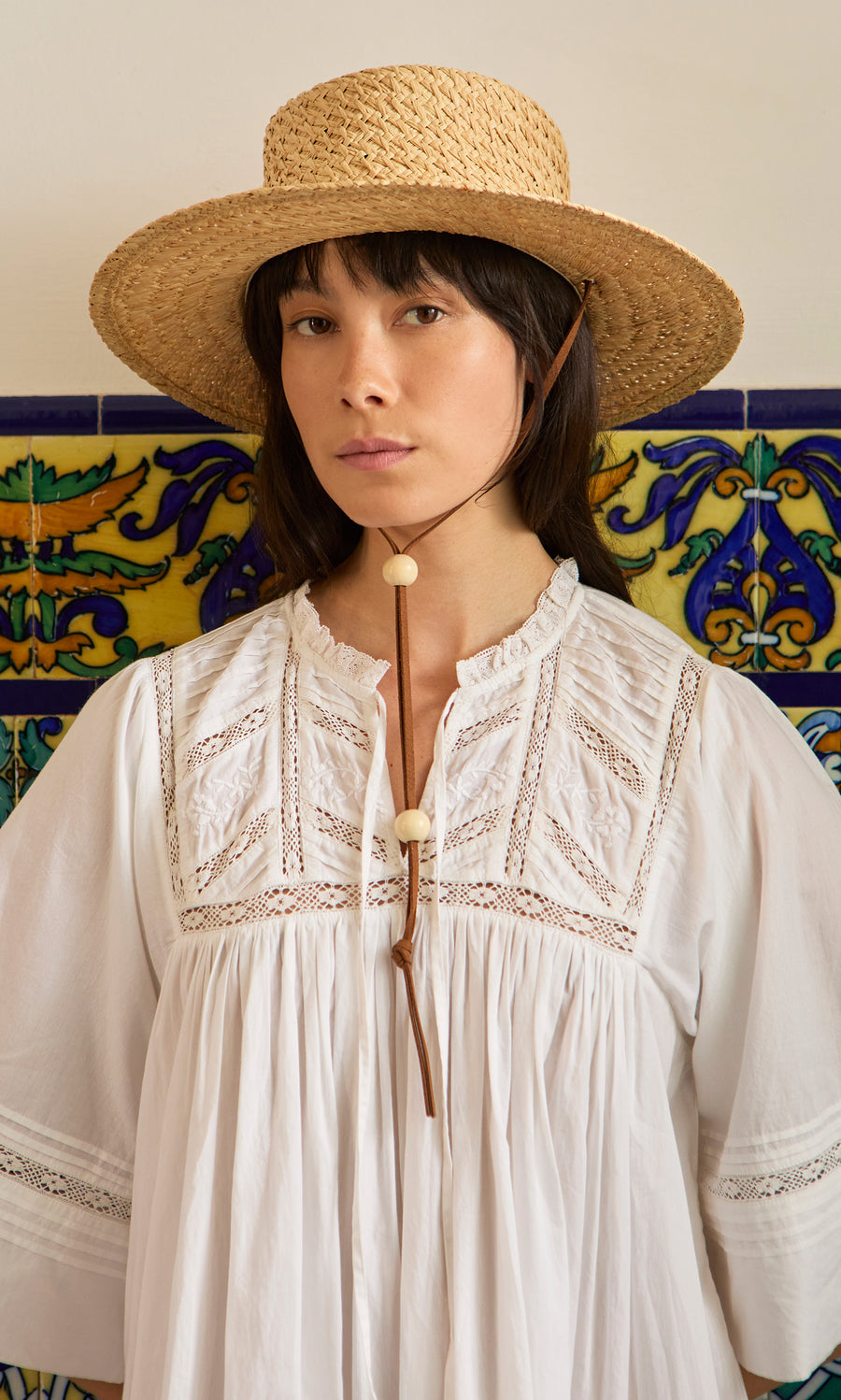 Plümo  Woman in white dress wears a Riva Straw Hat, standing before a colorful, patterned tiled wall.