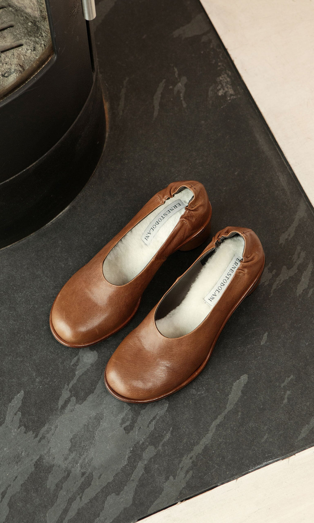 Plümo- A pair of Solene Heels in brown leather rests on a dark stone floor near a black metal fixture.