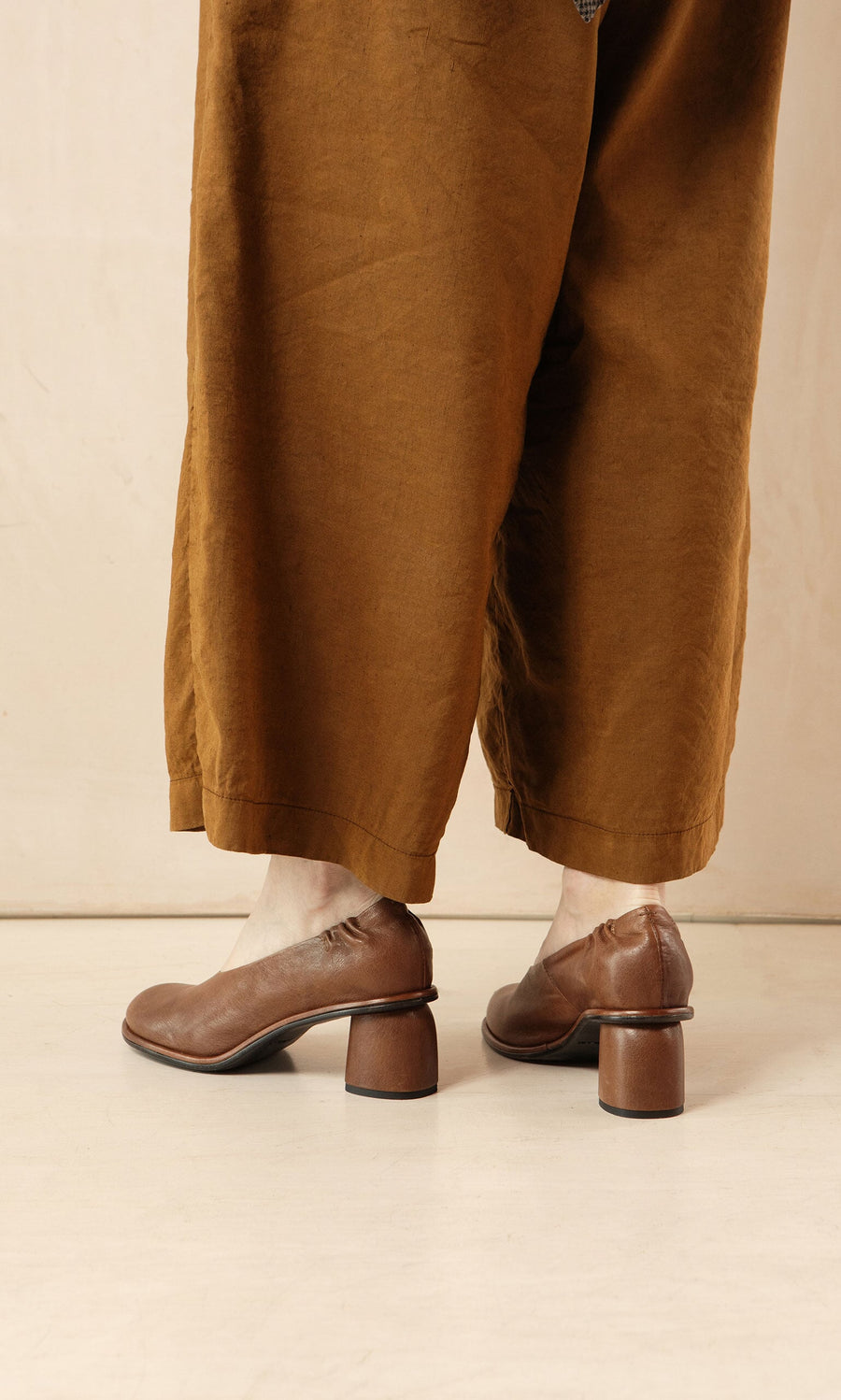 Plümo- Person standing on a light floor against a beige wall, wearing wide brown pants and Solene Heels.