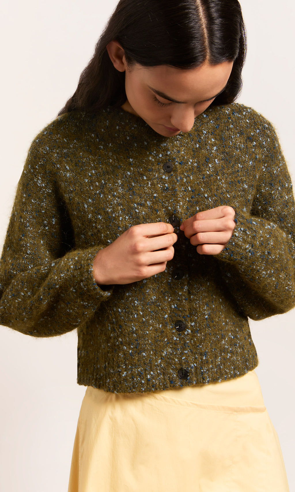 Plümo  Wearing the Fir Cardigan, they button up over a light yellow skirt, gazing down at their hands.