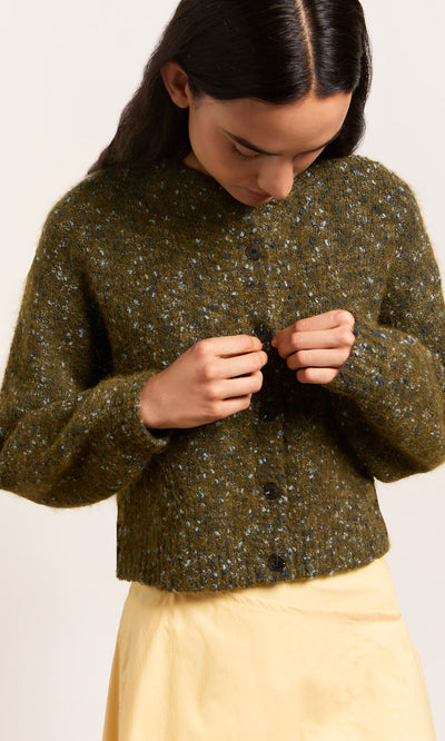 Plümo  Wearing the Fir Cardigan, they button up over a light yellow skirt, gazing down at their hands.
