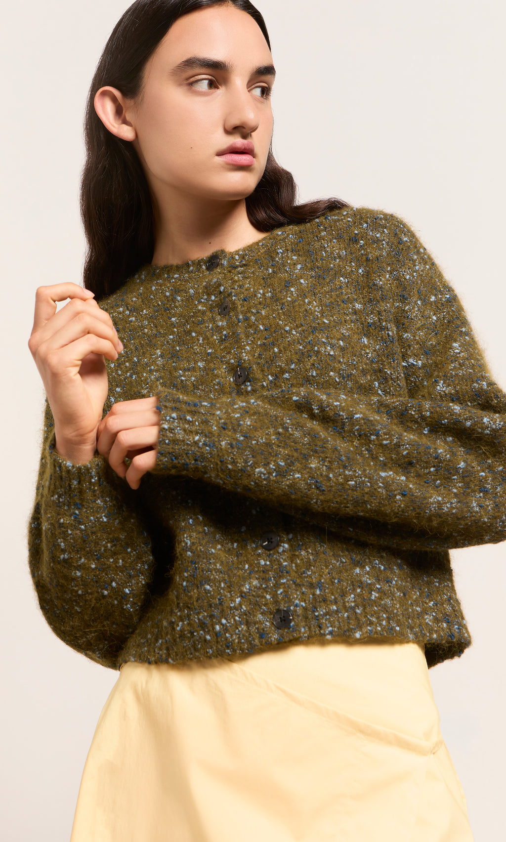 Plümo  Model in the Fir Cardigan and a yellow skirt, looking left against a simple backdrop.
