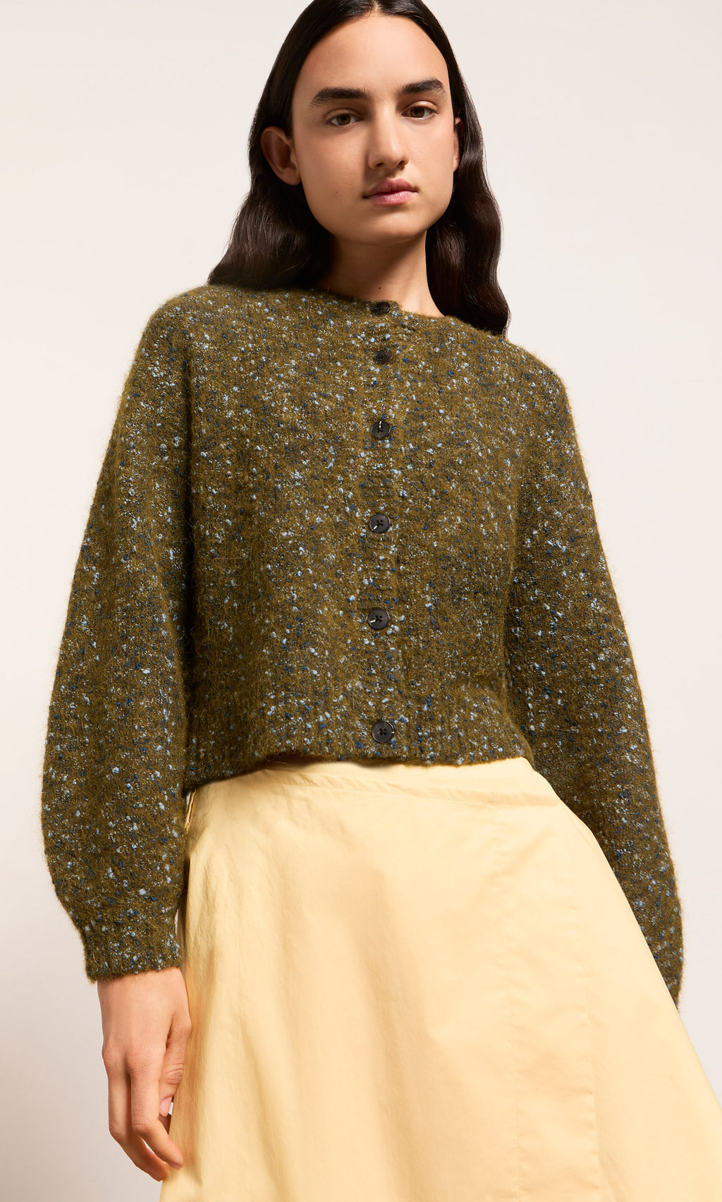 Plümo  Person in a green Fir Cardigan and light yellow skirt, standing against a plain background.