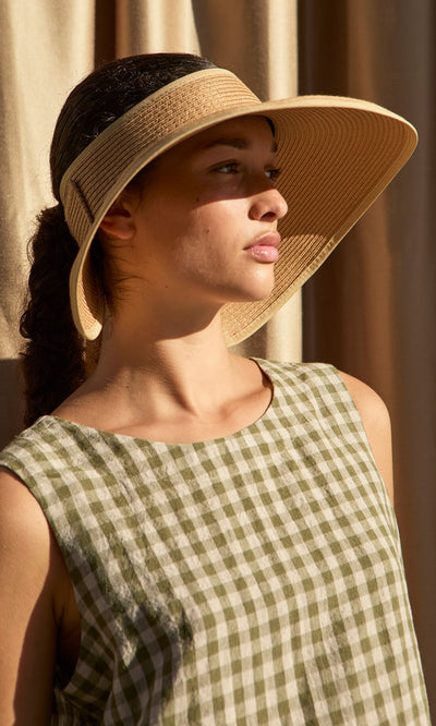 Plümo- Woman in a green gingham dress stands in sunlight, wearing the Miami Sun Visor and looking to the side.