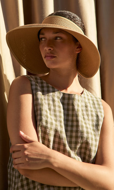 Plümo- Woman wears the Miami Sun Visor and a plaid sleeveless dress, standing with arms crossed in sunlight.