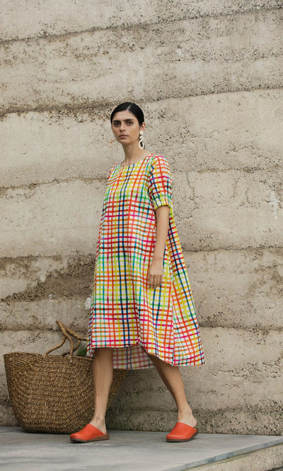 Plümo- Woman in the hand-dyed Thea Dress with orange shoes stands near a woven bag by a textured beige wall.