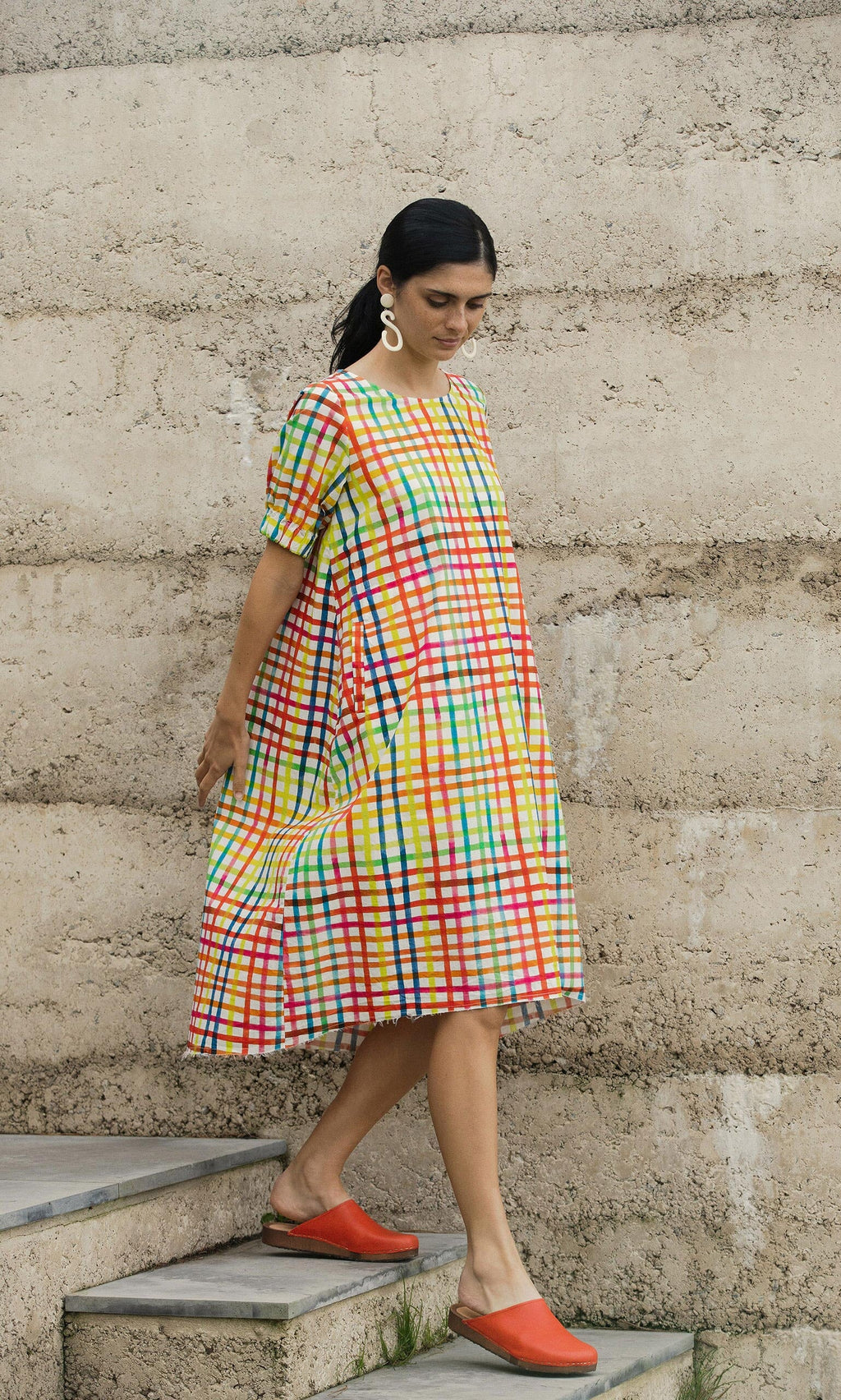 Plümo- Woman in the Thea Dress and red shoes walks down steps against a textured concrete wall.