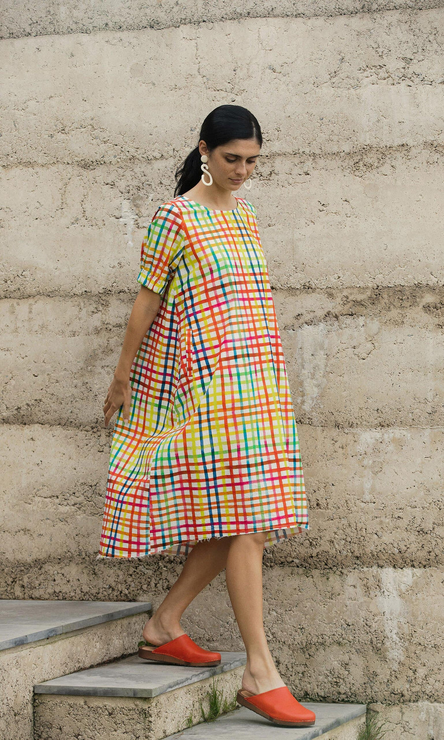 Plümo- Woman in the Thea Dress and red shoes walks down steps against a textured concrete wall.